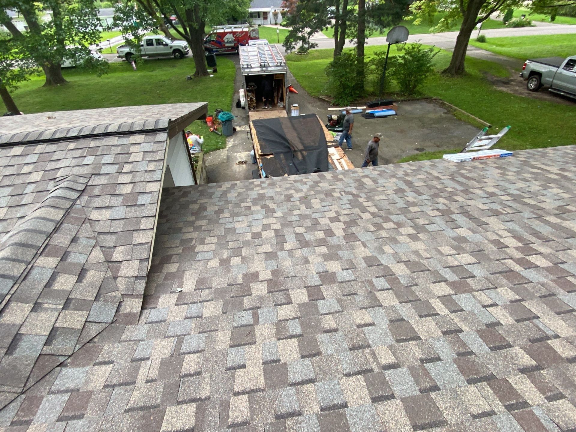 Roof shingles with workers and a dumpster in a tree-lined residential yard below