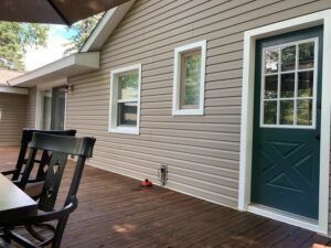 Covered porch with brown siding, teal door, white-trim windows, and wooden deck with black chair.