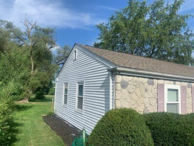 Small house with gray siding, stone wall, and green shrubs beside a grassy yard under a blue sky