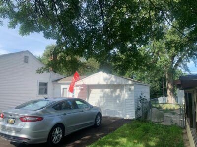 Silver car parked in a driveway beside a white garage, with a red flag and leafy tree overhead.