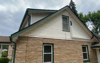 Two-story house with tan siding, stone lower facade, and green trim under a cloudy sky