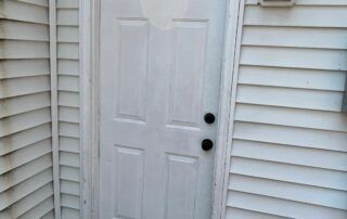 White exterior door with two black knobs, set in beige vinyl siding.