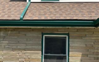 Close-up of a tan brick house exterior with a brown shingled roof and green trim around a window.