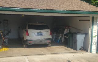 Car parked in a cluttered open garage with bins and tools on both sides.