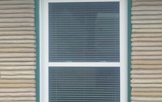 White-framed window with closed blinds on a beige-sided house exterior