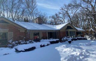 Snow-covered single-story house with a front porch and bare winter trees
