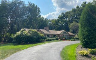 Curved driveway leading to a tan house surrounded by lush green trees and lawn under a blue sky