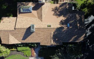 Aerial view of a brown shingled house roof with skylights, vents, and trees casting shadows.