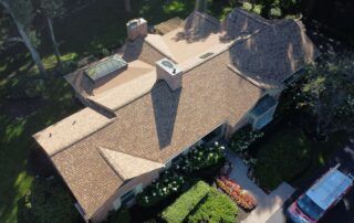 Aerial view of a brown-roofed house surrounded by trees and a driveway with cars nearby.