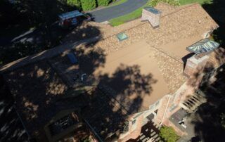 Aerial view of a tan shingled house roof with skylights and tree shadows.