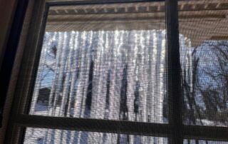 Icicles hanging from a roof edge outside a window with bare winter trees in the background