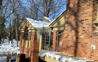 Snow-covered brick house exterior with large windows and leafless trees in winter