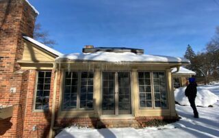 Snow-covered sunroom with large windows beside a brick house; person walking in the yard