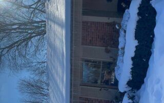 Snow-covered brick house with a window and trees in winter.