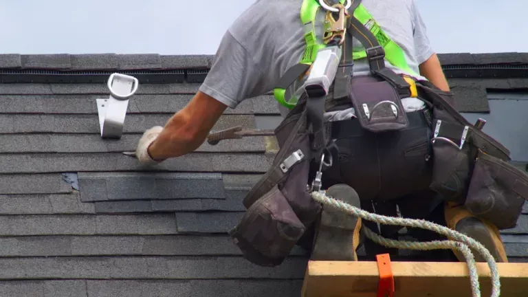 Worker on a roof installing dark shingles, wearing safety harness and tool belt.