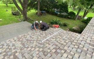 Workers repairing roof shingles beside a green garden, with tools and materials on a sloped rooftop.
