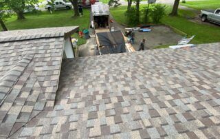 View of a shingled roof overlooking a wet walkway and green yard with trees and outdoor equipment