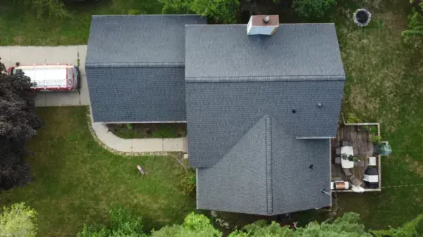 Aerial view of a gray-roofed house with a small patio and green yard, next to a driveway and parked trailer
