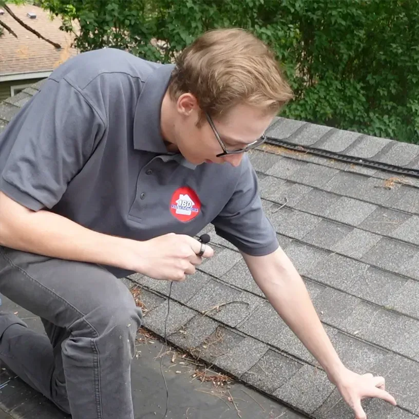 Person kneeling on a shingled roof, inspecting a section with a tool near a vent opening.