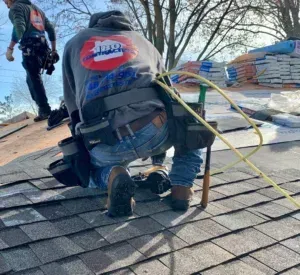 Roofers working on a shingled roof, one kneeling near safety ropes and stacked materials.