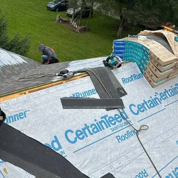 Roofing workers installing CertainTeed shingles on a steep gray roof with stacked materials nearby