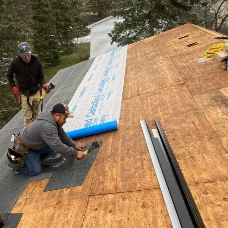 Two workers installing roofing underlayment on a sloped roof, with shingles and black trim nearby.