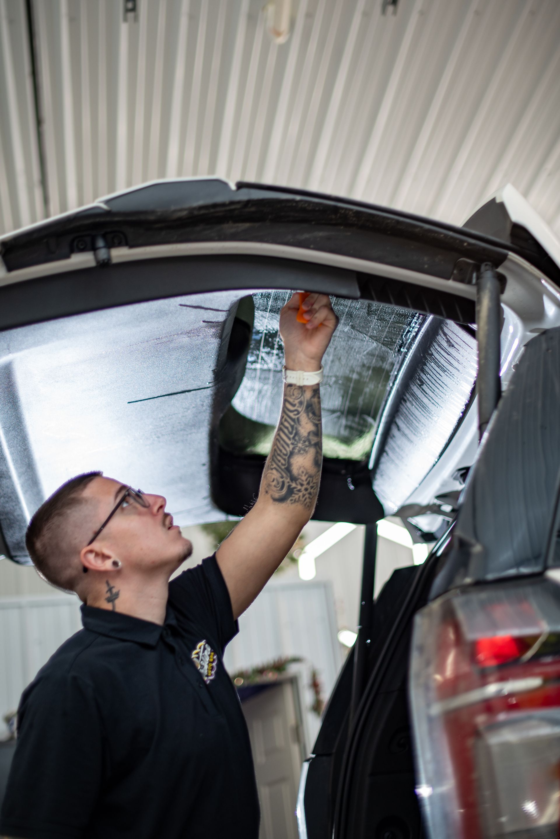 Person applying tint to a car window. They are wearing glasses and have arm tattoos. In a garage setting.
