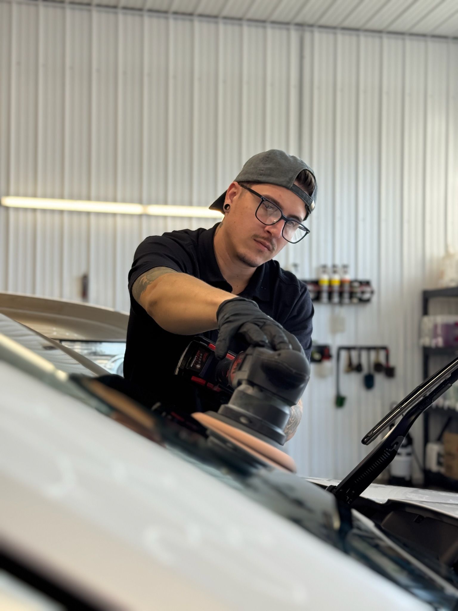 Man in a cap and glasses polishes a car windshield in a well-lit garage.