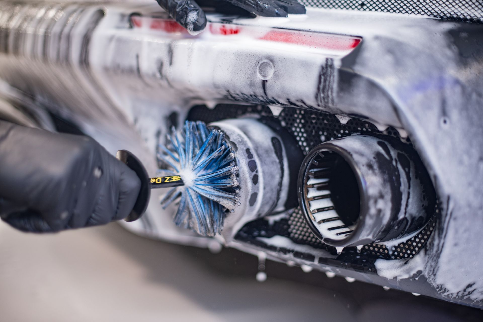 Gloved hand scrubs a car exhaust pipe with a brush, covered in white soap suds.