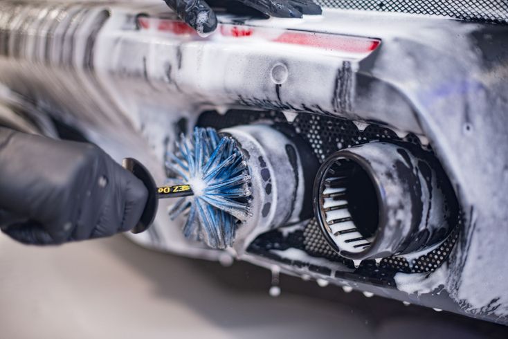 Gloved hand scrubs a car exhaust pipe with a brush, covered in white soap suds.