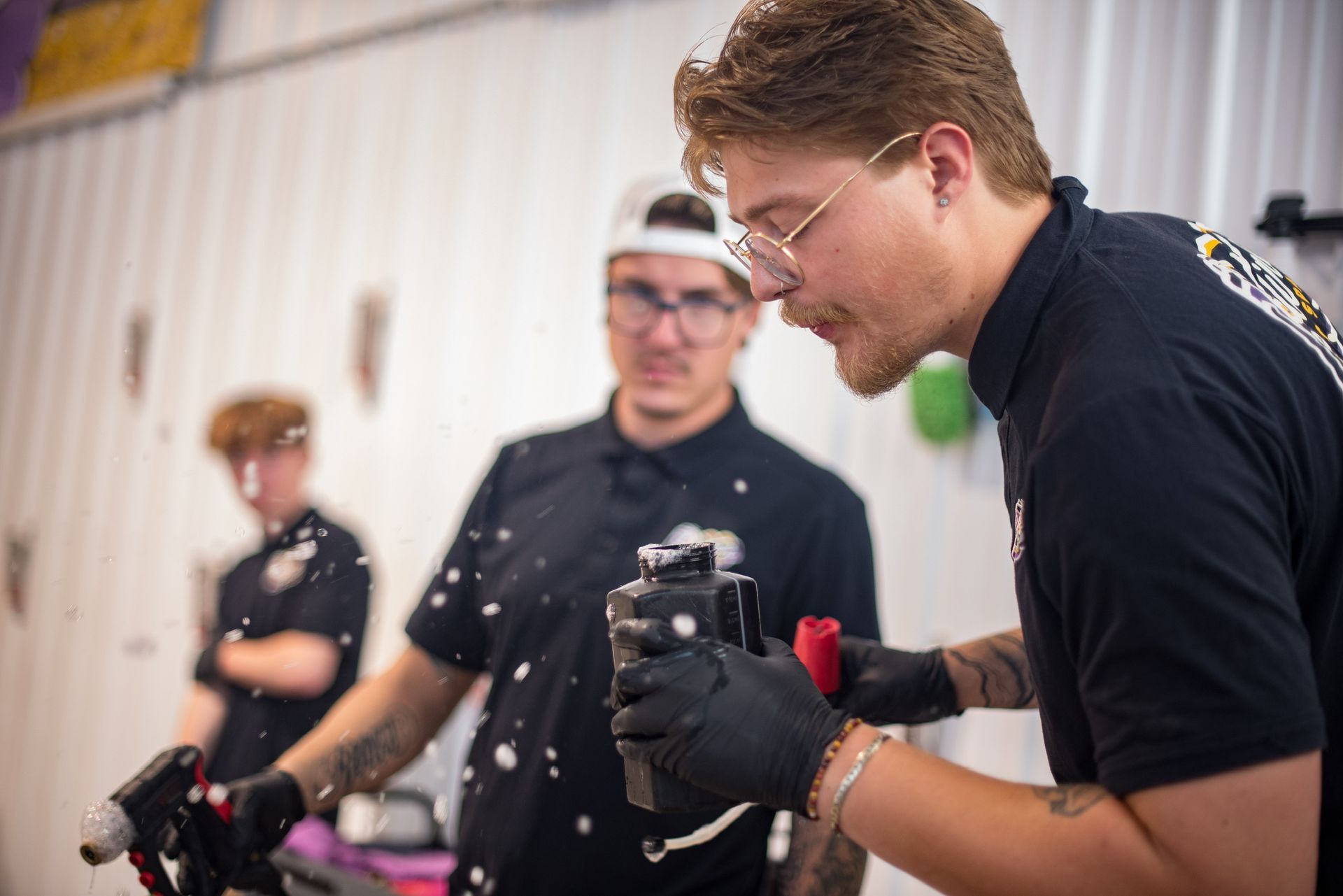 Three people in a workshop; one spraying, others watching. Paint splatters on the person spraying and a shirt.