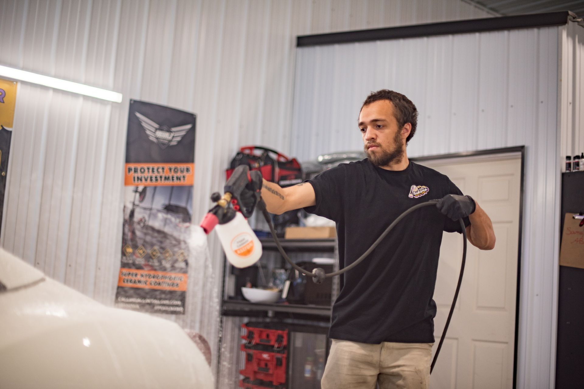 Man spraying foam onto a white car inside a shop. He wears a black shirt, gloves, and holds a foam cannon.