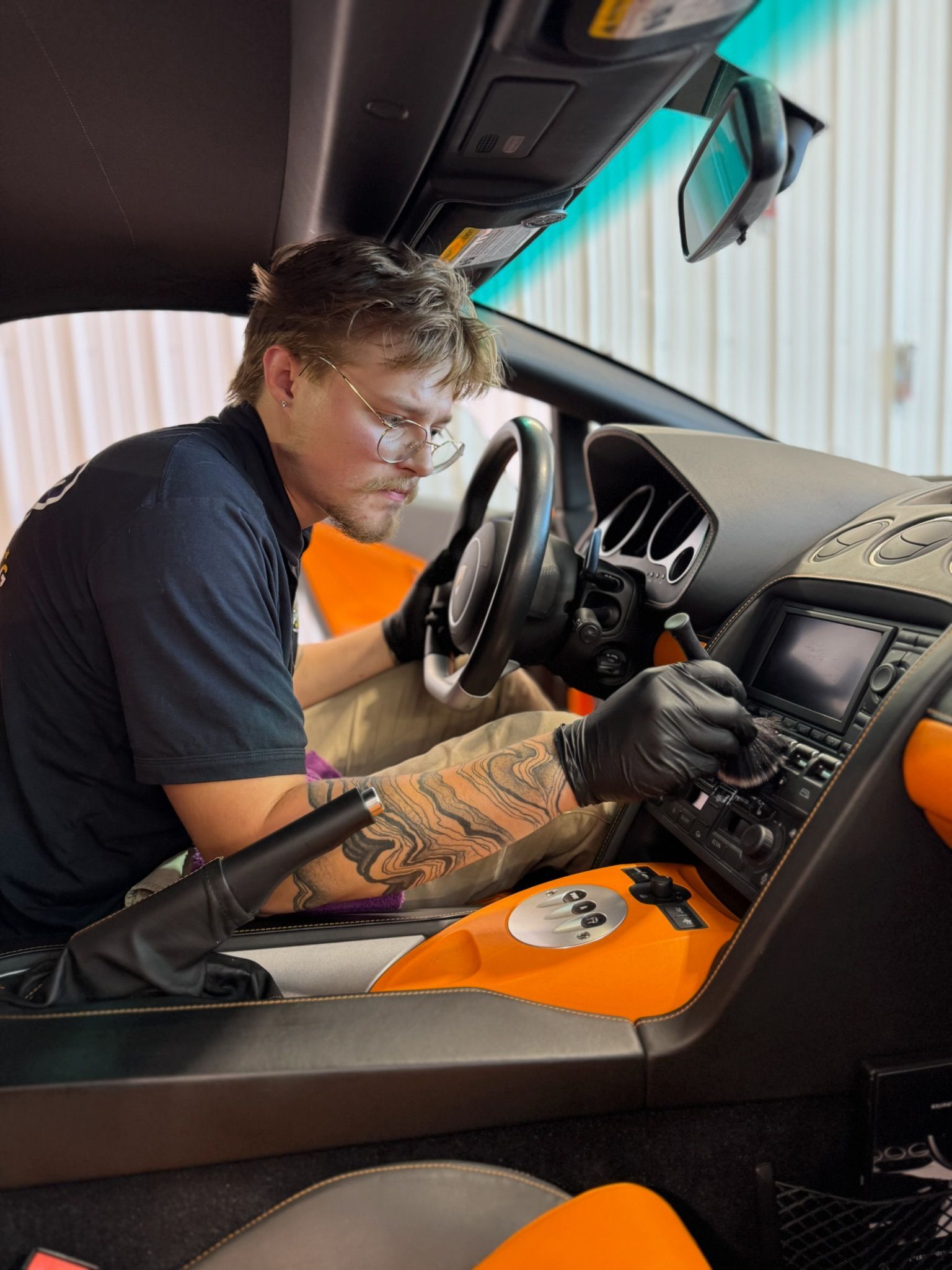 Man cleaning a car dashboard with a brush. He wears gloves, glasses, is in an orange-accented car.