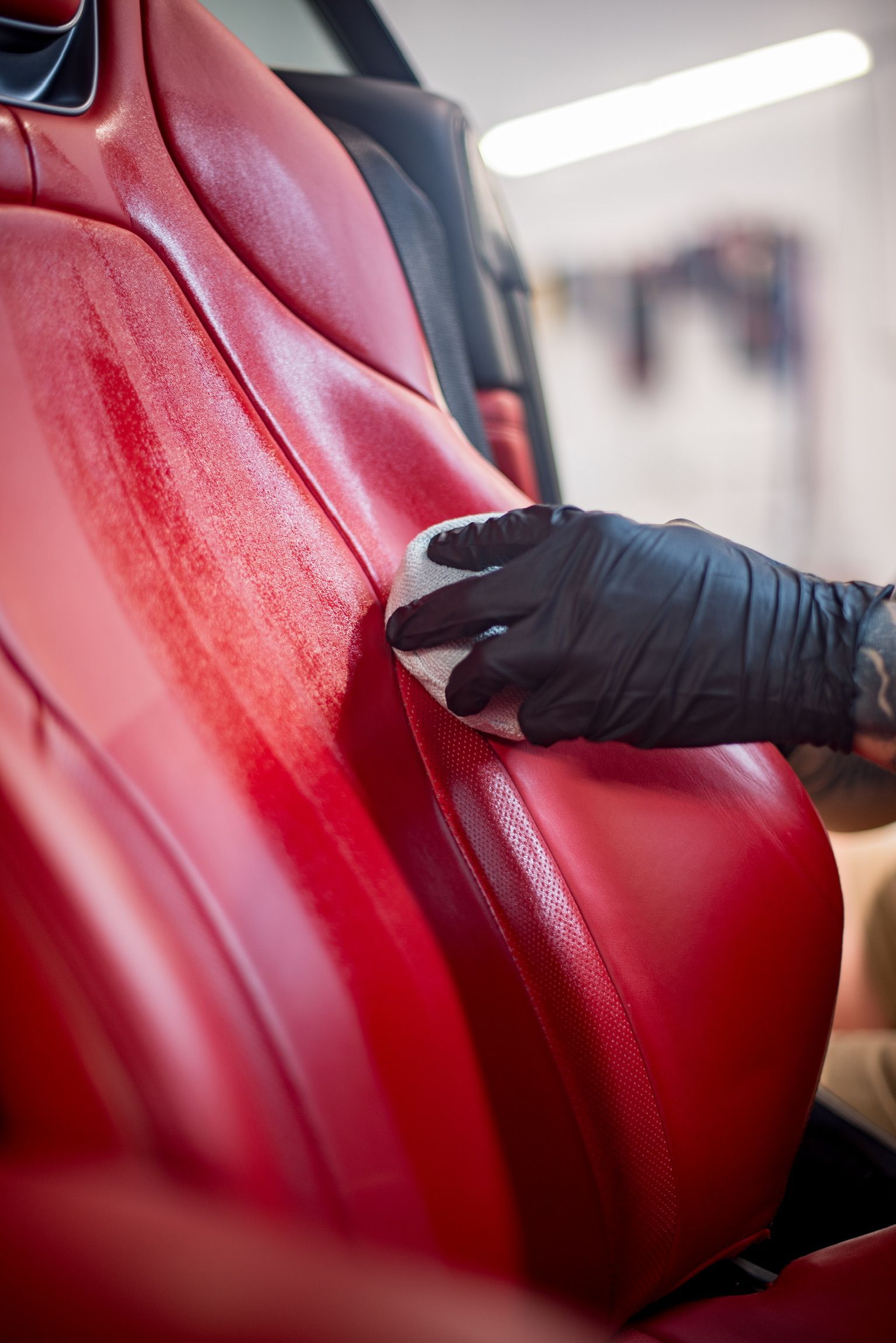 Person in black glove cleaning red leather car seat with a white cloth.