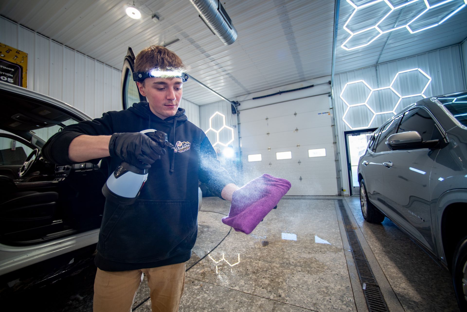 Person spraying a purple cloth with liquid in a garage. They are wearing a headlamp, gloves, and hoodie. A car is visible.
