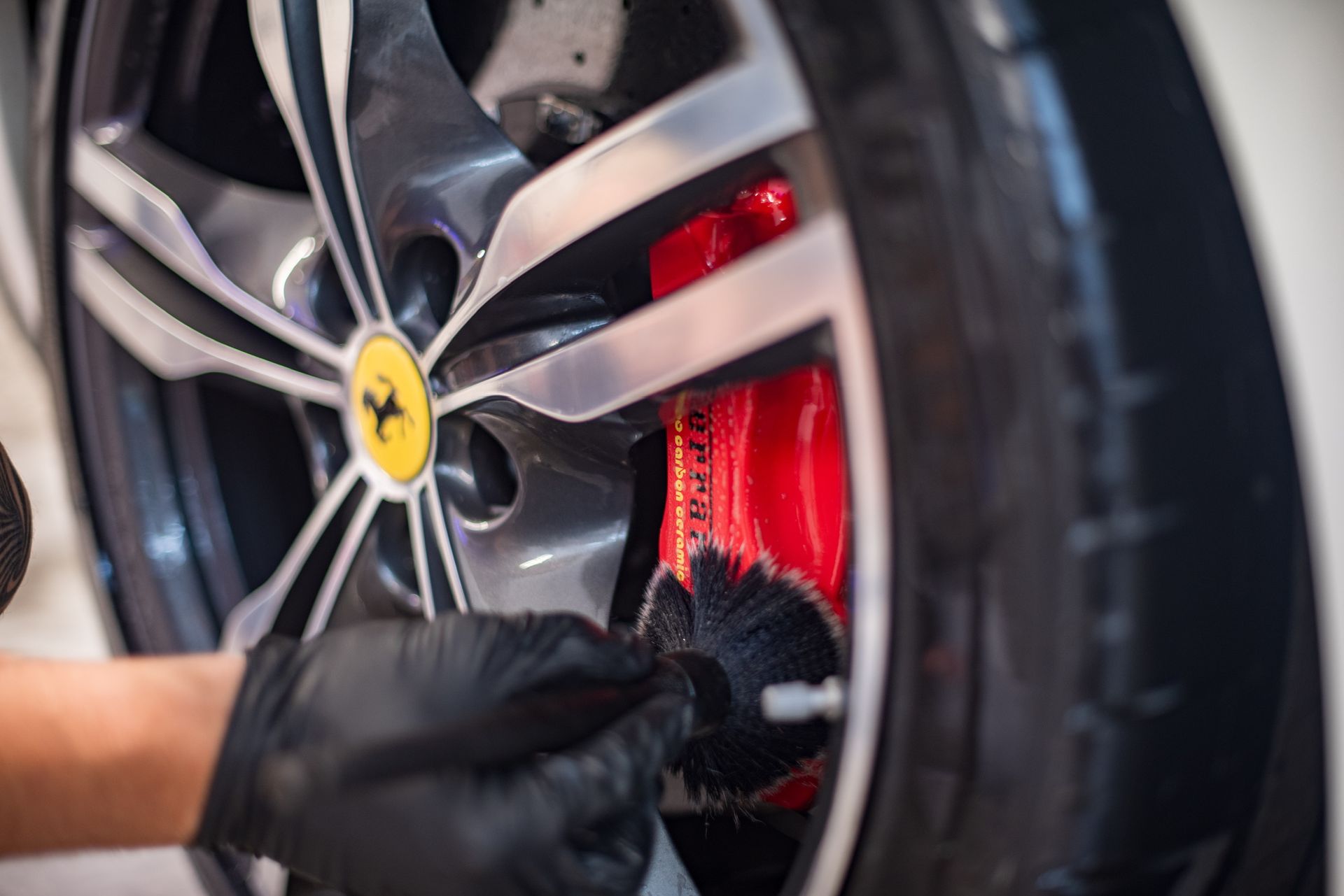 Black-gloved hand cleaning a red Ferrari brake caliper with a brush, wheel in background.