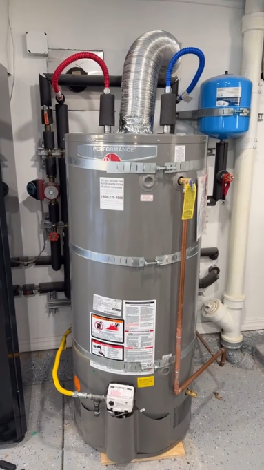 A large gray water heater is sitting in a room next to pipes.