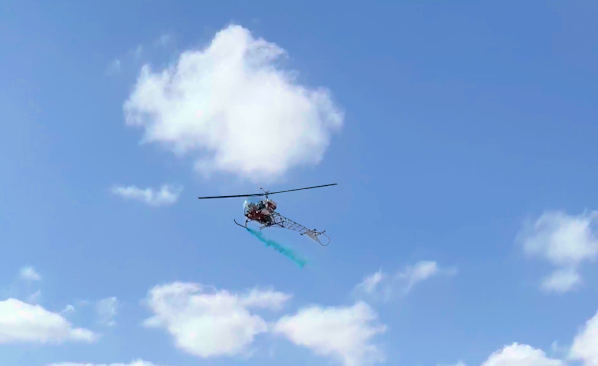 Helicopter flying in a blue sky with fluffy white clouds. The helicopter has a light blue trail.