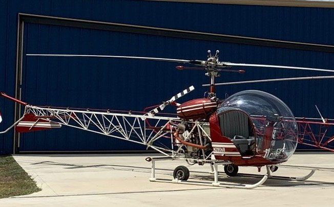Red and white Hiller UH-12E helicopter with a bubble cockpit parked on a tarmac in front of a blue hangar.