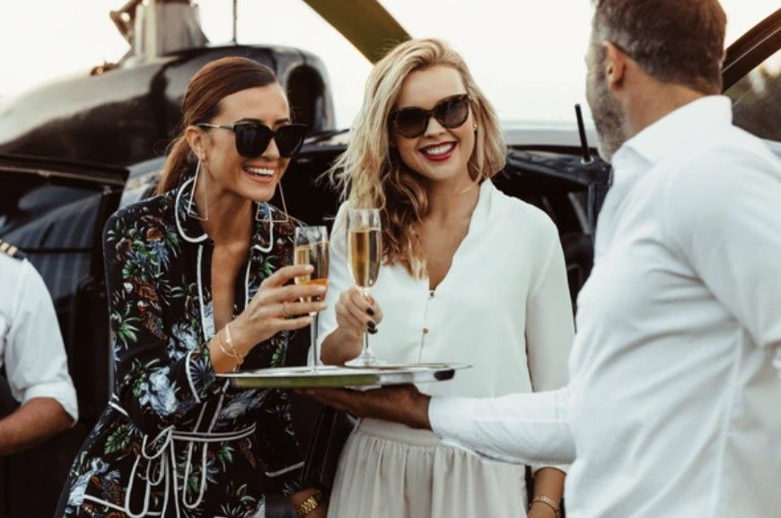 Two stylish women in sunglasses cheers champagne glasses with a waiter near a helicopter; outdoor setting.