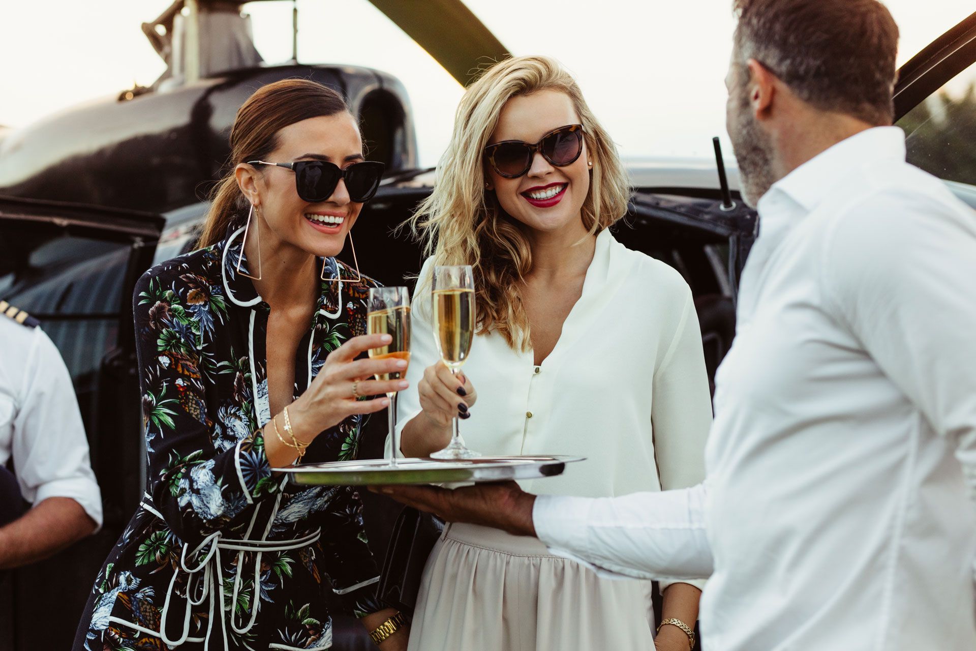 Two stylish women in sunglasses cheers champagne glasses with a waiter near a helicopter; outdoor setting.