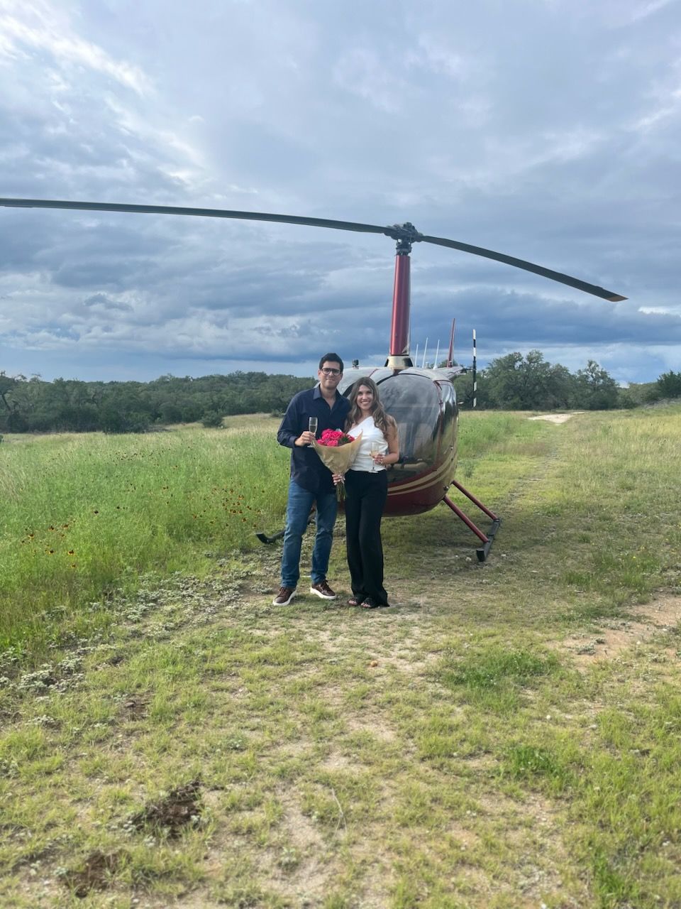 Couple standing in front of a red helicopter, holding flowers and champagne, on a grassy field. Cloudy sky in background.