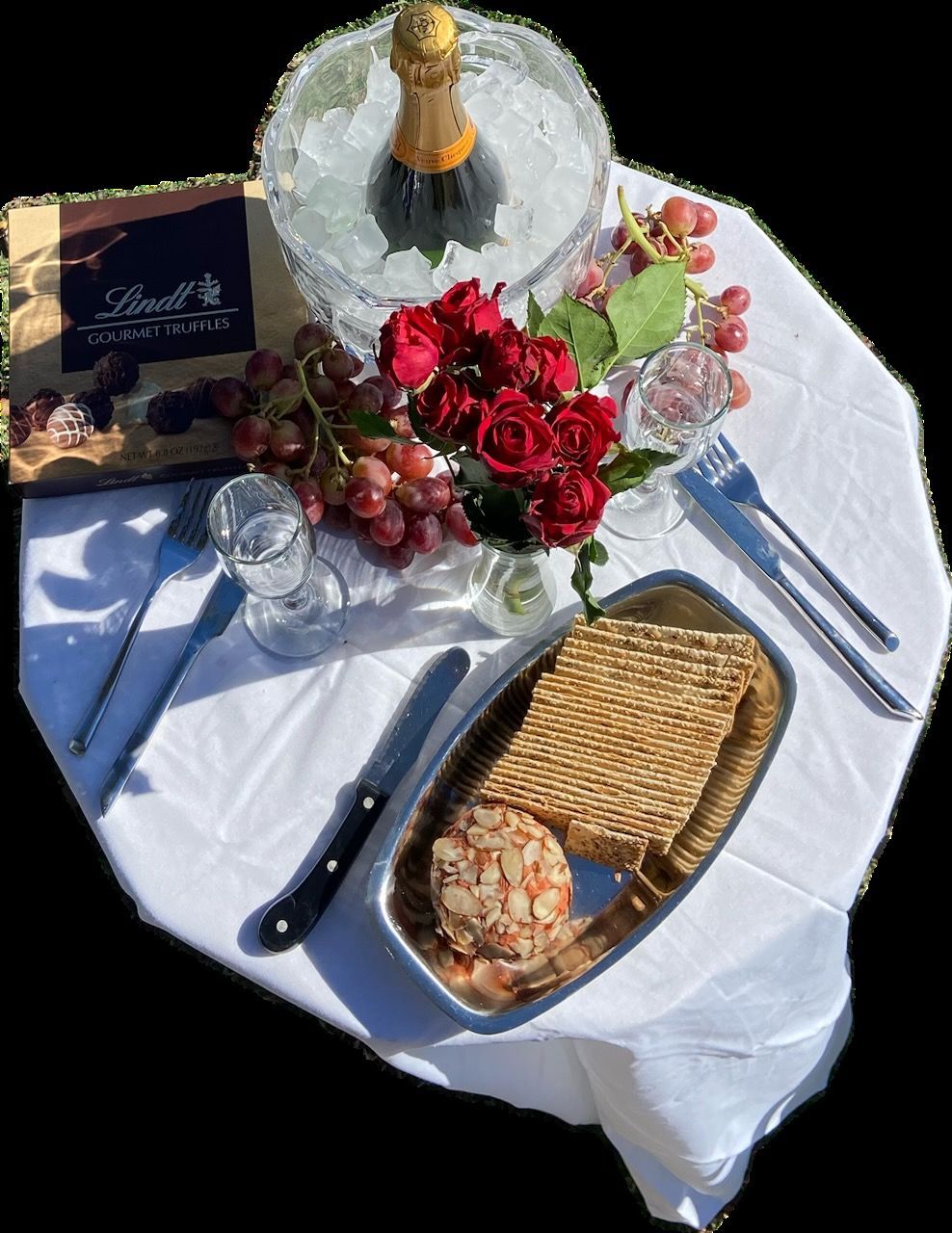 Picnic setup with champagne, roses, grapes, crackers, cheese, and chocolates on a white tablecloth.