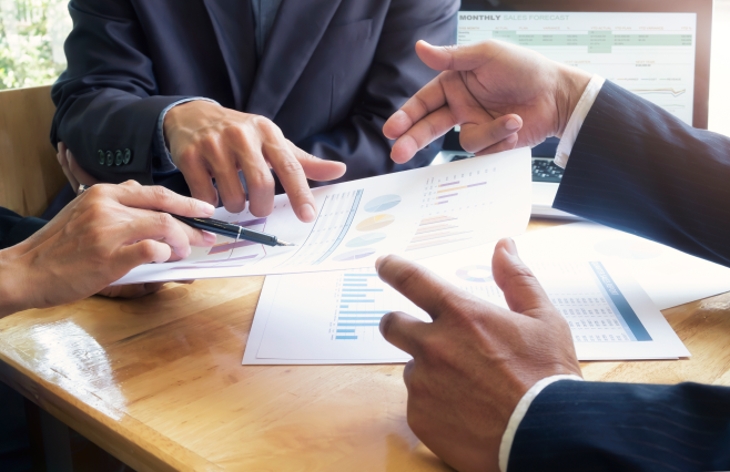 Business professionals in suits reviewing financial documents, pointing and discussing at a wooden table.