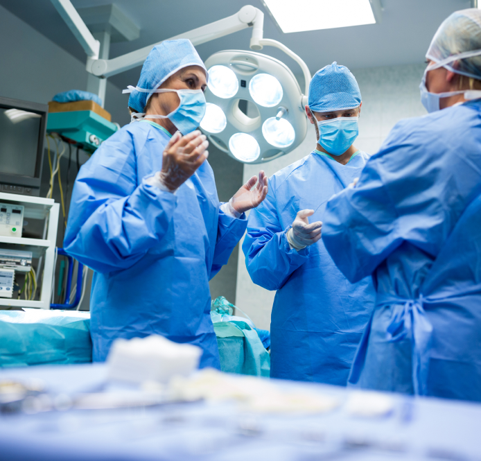 Medical team in blue scrubs and masks in an operating room, discussing a procedure under bright lights.