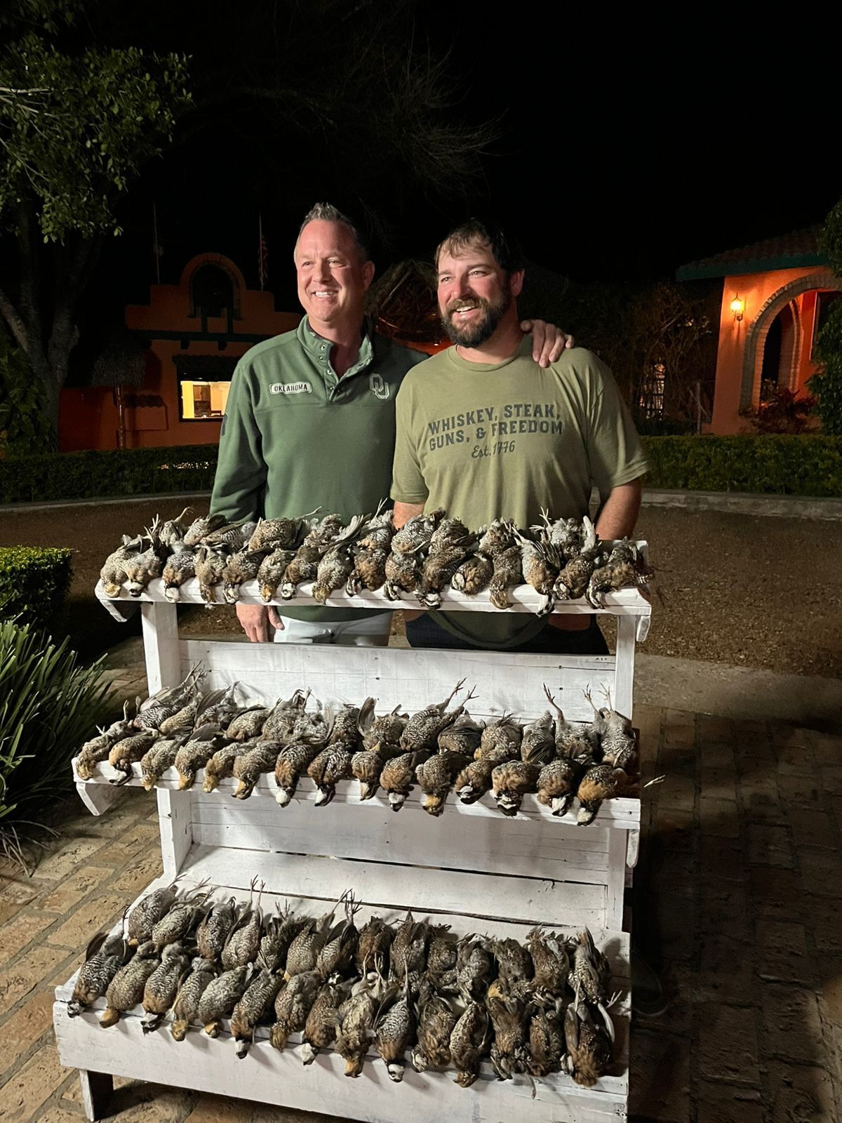 Two men pose behind racks of ducks at night outdoors