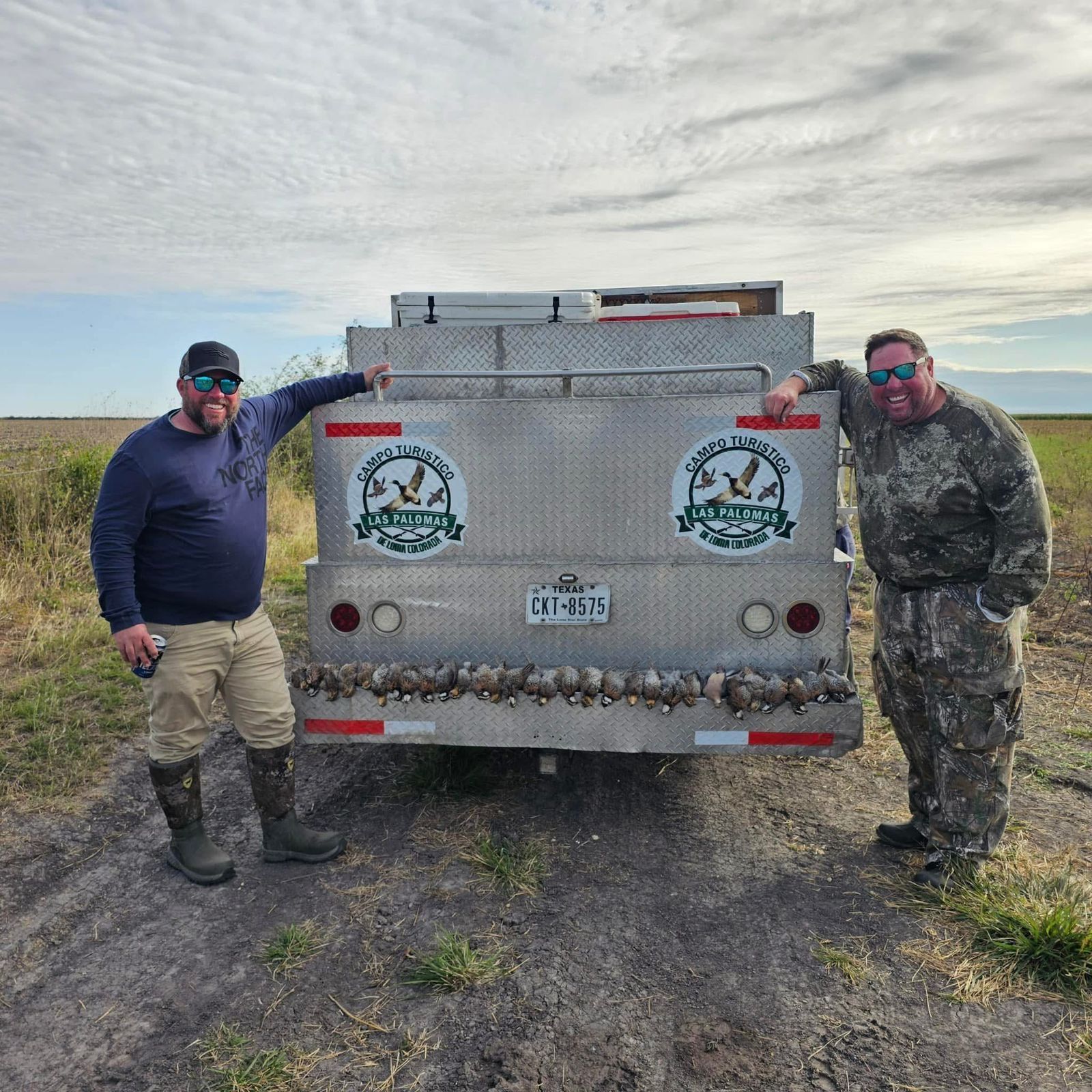 Two camouflaged people posing beside a silver utility trailer in a grassy field under a cloudy sky