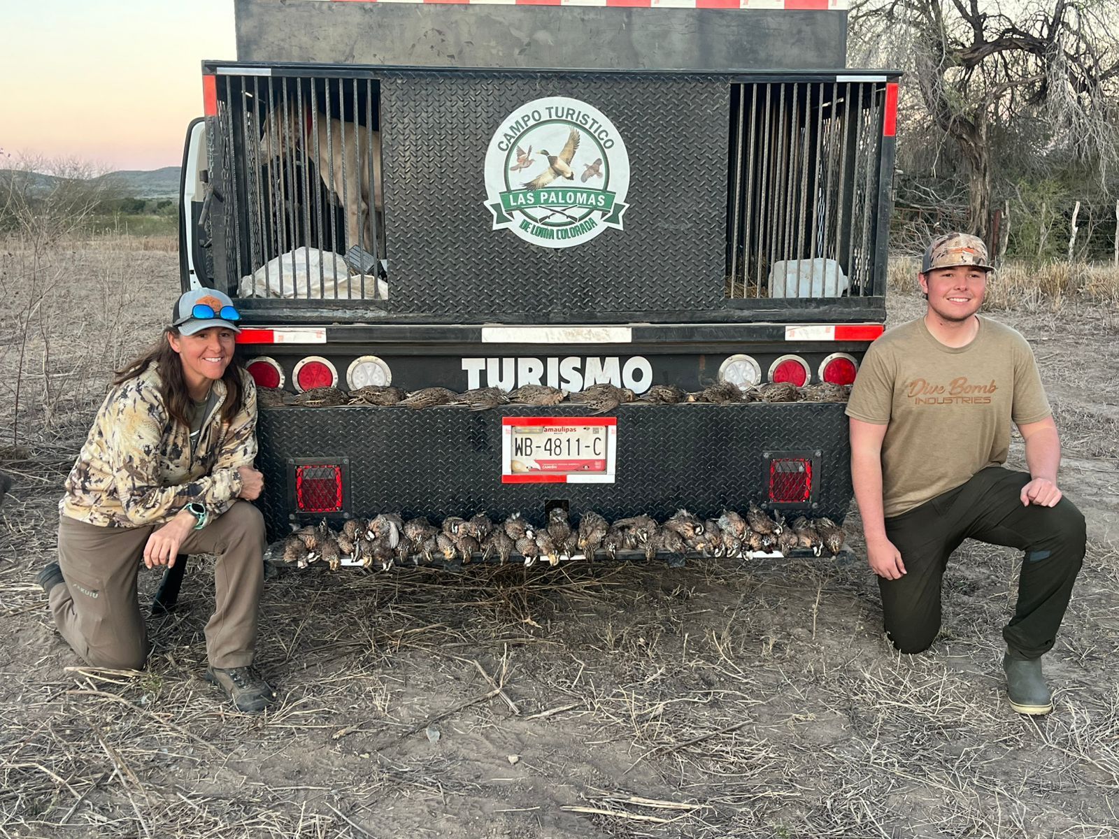 Two people kneel beside a black off-road vehicle with a “TURISMO” sign on the back.