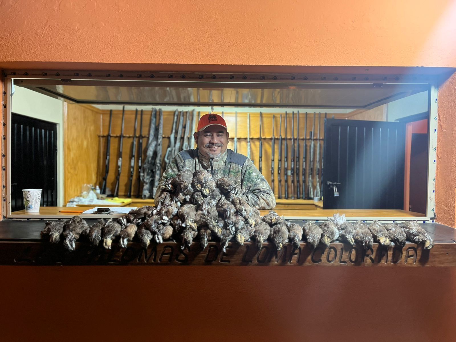 Hunter standing behind a counter with a row of hanging game birds and rifles in the background