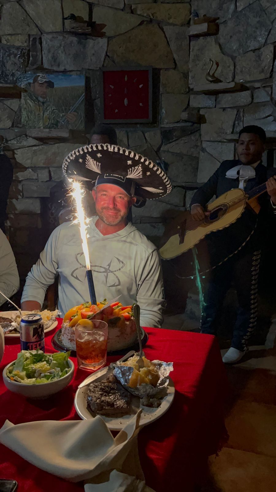 Dinner table with lit birthday cake and a man wearing a sombrero, with a guitarist in the background.